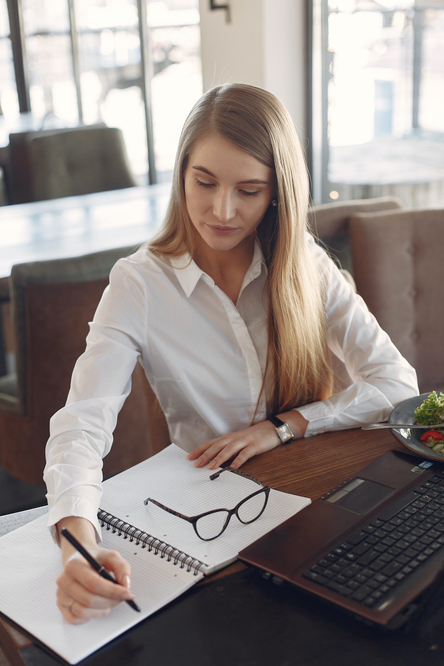 young-woman-writing-in-notebook-while-working-on-laptop-3874629